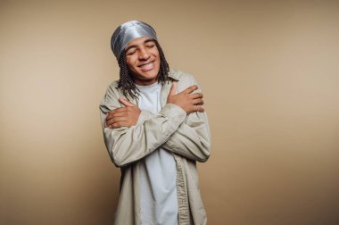 Gümüş renkli başörtüsü olan mutlu genç bir adamın portresi kollarını çaprazlıyor, sıcak bir şekilde gülümsüyor, bej arka planda kendini sevme duygusunu resmediyor.