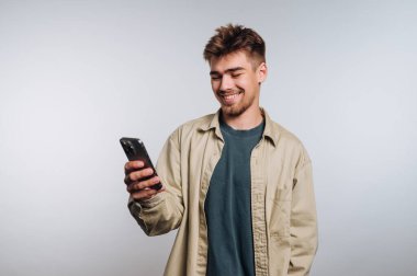 Genç adamın portresi, beyaz arka planda mutluluğu ve zevki ifade eden geniş bir gülümseme gösteren akıllı telefonuyla neşeyle meşgul.