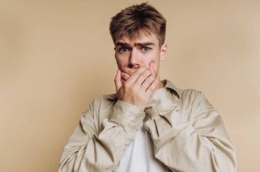 Şaşırmış genç adamın portresi, bej arka plandaki şaşkınlık anını yansıtan bir şok ile ağzını kapatıyor.