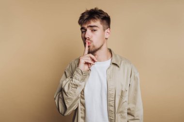 Genç bir adamın portresi sessizlik için bir jest yapar, bej arka plan üzerinde bir anlık tefekkür gösterir.