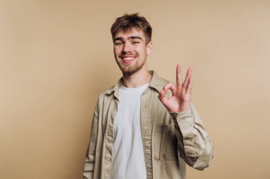 Genç, neşeli bir adamın portresi gülümsüyor ve iyi bir jest yapıyor, bej arka planda olumlu ve olumlu bir duyguyu kolayca aktarıyor.