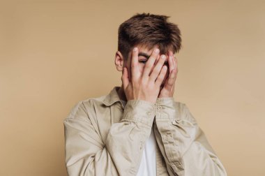 Genç adamın portresi, ellerini bej arka planda sakin bir atmosferde şakacı bir şekilde gizliyor.