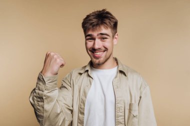 Mutlu genç bir adamın portresi yüzünde sıcacık bir gülümsemeyle kutlamada yumruğunu kaldırıyor, bej arka planda pozitiflik ve neşe yayıyor.