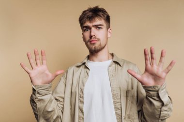 Genç bir adamın portresi ya savunma jesti olarak ellerini kaldırır ya da bej arka planda durur.