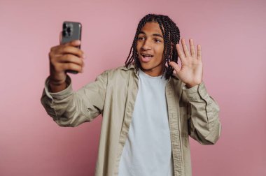 Genç adamın portresi mutlu bir şekilde akıllı telefonlarına el sallıyor, pembe arka planda dostça bir etkileşimi somutlaştırıyor.