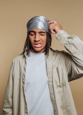 Örgülü saçlı genç bir adamın portresi bej arka planda kafasını kaşıyarak kafa karışıklığını ve hayal kırıklığını ifade eder.