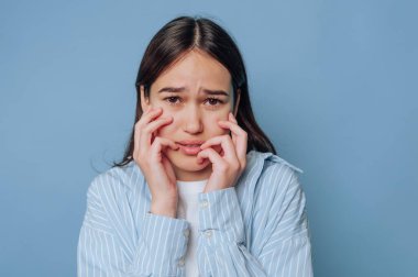 Genç bir kadın iki eliyle yüzünü tutarak endişe ve endişe gösterir. Uzun saçları var ve çizgili bir gömlek giyiyor. Arkaplan düz mavi, ifadesini vurguluyor..
