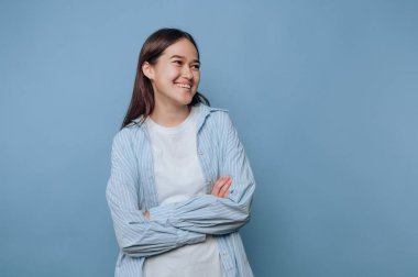 Genç bir kadın kollarını kavuşturmuş, sıcak bir şekilde gülümsüyor. Açık mavi bir gömlek ve sade beyaz bir bluz giyiyor. Sıcak bir yerde samimi ve cana yakın bir hava yaratıyor..