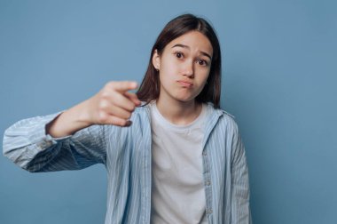 Genç bir kadın, saf mavi bir arkaplan önünde durup yüzünde düşünceli bir bakışla parmak işareti yaparak merakını ya da düşüncenin yansıtmasını yapar..