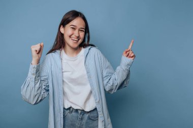 Genç bir kadın gülümseyerek ve kolları havada dans ederek neşe ifade eder. Açık mavi bir gömlek ve kot pantolon da dahil olmak üzere sıradan bir kıyafet giyiyor..