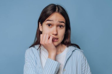 Genç bir kadın, mavi arka planlı sakin bir yerde tırnaklarını yerken endişeli bir bakış sergiliyor. Onun ifadesi gerginliği ve endişeyi yansıtıyor..