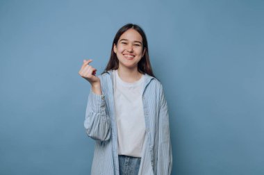Neşeli genç bir kadın açık mavi bir arka planın önünde durup parmaklarıyla kalp hareketi yapıyor. O gündelik bir kıyafet giyiyor ve uzun saçları var. Sahne pozitifliği ifade eder.