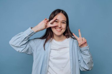 Genç bir kadın mavi bir arkaplanda duruyor, iki eliyle barış işareti yaparken ışıl ışıl gülümsüyor. Günlük kıyafetleri ve şakacı ifadesi mutluluk ve güven veriyor..