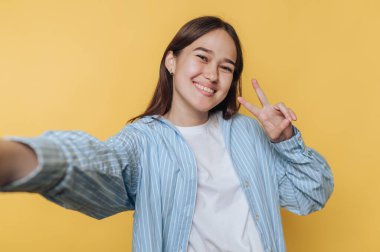 Genç bir kız selfie çekerken mutlu bir şekilde gülümsüyor. O, açık mavi çizgili bir ceket giymiş, açık sarı bir zemin üzerinde, bir barış işareti gösteriyor..