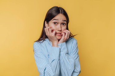 Genç bir kadın tırnaklarını yerken ve kameraya bakarken gergin bir yüz ifadesi sergiliyor. Açık mavi çizgili bir gömlek giyiyor, parlak sarı bir arka plana karşı..