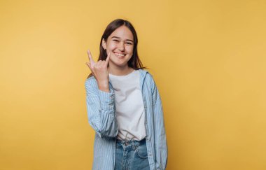 Genç bir kadın canlı sarı bir arka planın önünde duruyor, geniş bir gülümseme ve gelişigüzel giyinmiş bir şekilde el hareketi gösteriyor. Sahne bir neşe ve pozitiflik hissi veriyor..