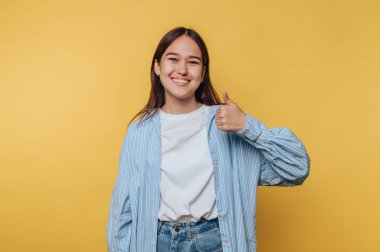 Genç bir kadın parlak sarı bir arkaplanın önünde duruyor. Gülümsüyor ve olumlu ve mutlu bir ruh halini temsil eden baş parmağını kaldırıyor..