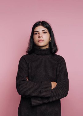 Genç bir kadın kollarını kavuşturmuş, siyah bir kazak giyiyor. Yumuşak pembe bir arka plana karşı ciddi bir ifade sergiliyor, eşsiz tarzını sergiliyor..