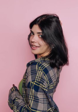 Genç bir kadın, ekose gömlek giymiş, neşeli bir ifadeyle kendinden emin bir şekilde ayakta duruyor. Canlı pembe arkaplan onun hareketli tavrını ve tarzını arttırıyor..