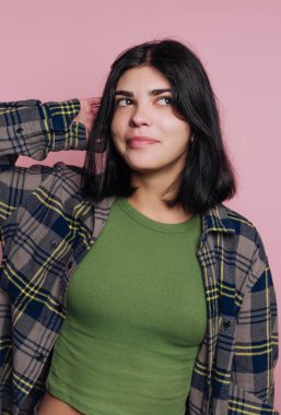 Bir kadın yeşil bluz ve ekose gömlek giyerek pembe bir arkaplanda duruyor. Yüzü şen şakrak ve rahat, saçlarına dokunurken kendine güveni tam..