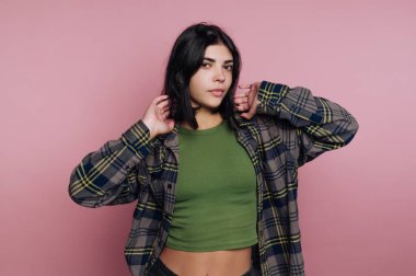 Genç bir kadın elleri başının üzerinde duruyor, yeşil bir bluzun üzerine ekose bir gömlek giyiyor. Pembe bir arkaplana yaslanmış, kendine güvenen ifadesini ve günlük tarzını sergiliyor..