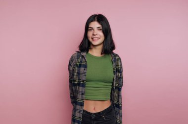 Genç bir kadın kendine güvenerek duruyor, şık bir elbiseyle gülümsüyor yeşil bluzu ve ekose gömleği var. Parlak pembe arka plan onun neşeli tavırlarına canlı bir dokunuş katıyor..