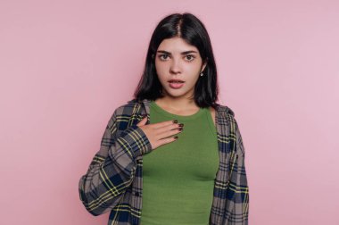 Genç bir kadın pembe bir duvarın önünde duruyor, şaşırmış görünüyor. Koyu renk saçları var ve ekose gömleğinin altında yeşil bir bluz giyiyor. Eli göğsünde, güçlü bir tepki gösteriyor..