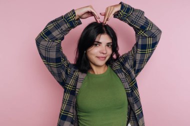 Genç bir kadın sevinçle elleriyle kalp şeklini oluşturuyor. Yeşil bir üst ve pembe bir duvarın önünde poz veren kareli bir ceket giyiyor. Gülümsemesi mutluluk ve güven gösteriyor..