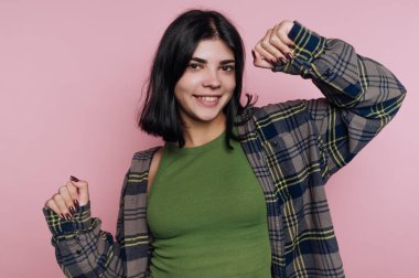 Genç bir kadın, yumuşak pembe bir arka plan önünde neşeyle gülüp dans ederken görülüyor. Yeşil bir üst katman giyiyor. Rahat bir ekose gömlek giyiyor. Eğlenceli ve kaygısız bir hava veriyor..