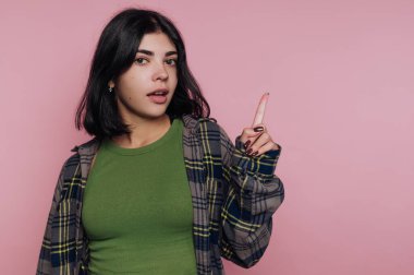 Genç bir kadın parlak pembe bir arka planın önünde duruyor, yeşil bir gömlek ve büyük bir ekose ceket giyiyor. Sanki bir şey ima ediyormuş ya da ima ediyormuş gibi parmağını dikkatle işaret ediyor..