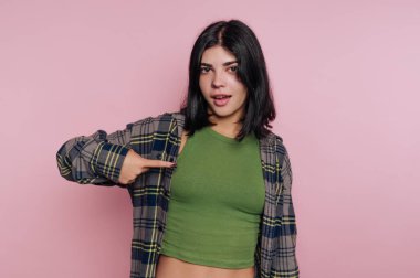 Genç bir kadın pembe bir arka plana karşı duruyor, yeşil bluz ve ekose gömlek giyiyor. Kendine güvenen bir gülümseme ile kıyafetini işaret ediyor..
