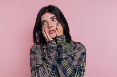 Genç bir kadın pembe bir arka plana karşı duruyor, kareli bir ceket giyiyor. Düşünceli bir yüz ifadesi sergiliyor, derin düşüncelere dalmışken ellerini yanaklarının üzerine koyuyor..