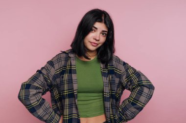 Genç bir kadın elleri kalçasında duruyor, yeşil bir bluzun üstüne ekose gömlek giyiyor. Pastel pembe bir arka plana karşı rahat ve kendinden emin bir ifade sergiliyor..