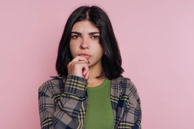 Genç bir kadın, düşünceli bir ifadeyle pembe bir arkaplanın önünde duruyor. Yeşil bir bluzun üzerine ekose bir ceket giyiyor ve derin düşüncelere dalmış gibi görünüyor..