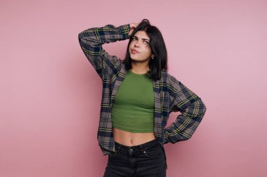 Genç bir kadın düşünceli bir ifadeyle ayakta duruyor, elleri başının üstünde. Yeşil bir üst ve açık fanila gömlek giyer, yumuşak pembe bir arkaplan karşı, düşünce önerir.