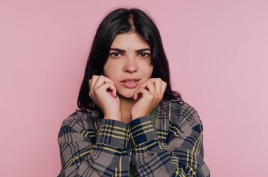 Genç bir kadın pembe bir arka planın önünde duruyor, ciddi bir ifadeyle doğrudan kameraya bakıyor. O, sıradan bir ekose gömlek giyiyor ve düşünceli bir şekilde yüzüne dokunuyor..