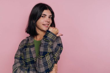 Genç bir kadın pembe bir duvarın önünde duruyor, gülümsüyor ve başparmağını kaldırıyor. Yeşil bir bluzun üzerine ekose gömlek giyiyor ve omuz hizasında siyah saçları var..