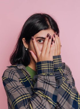 Pembe bir arka planın önünde duran genç bir kadın elleriyle yüzünü kapatıyor. Kareli bir ceket giyiyor ve uzun, manikürlü tırnaklarını gösteriyor, neşeli bir tavır sergiliyor..