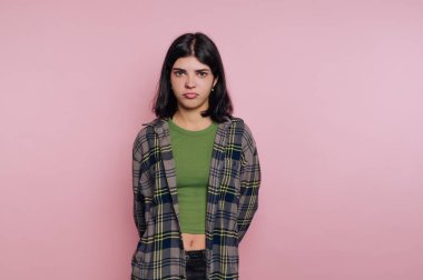 Omuz hizasında koyu saçlı genç bir kadın, sıradan bir kıyafetle ciddi bir şekilde poz veriyor. Bir stüdyo ortamında pembe bir zemine karşı duruyor, özgün bir anı yakalıyor..