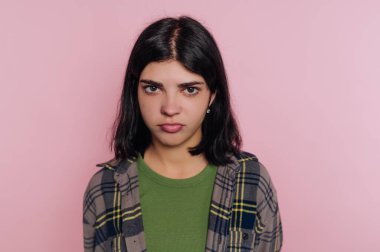 Genç bir kadın pembe bir arkaplan önünde tarafsız bir ifadeyle duruyor. Koyu renk saçları var, yeşil bluzunun üstüne ekose gömlek giyiyor ve doğrudan seyirciye bakıyor..