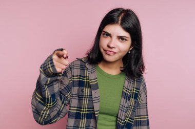 Genç bir kadın, neşeli bir sırıtışla, ekose bir ceketin altında yeşil bir gömlek giyerek güvenle ayakta duruyor. Seyirciyi neşeli bir bakışla işaret ediyor..