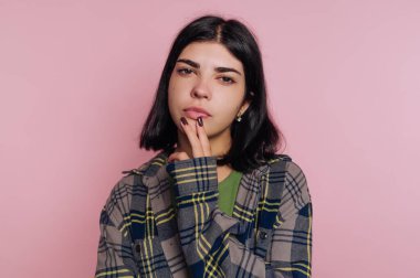 Genç bir kadın pembe bir duvarın önünde duruyor, düşüncelere dalmış. Ekose bir gömlek giyer ve elini dudaklarına koyup derin düşüncelere dalan bir yüz ifadesi gösterir..