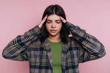 Genç bir kadın pembe bir duvarın önünde duruyor, parmaklarıyla şakaklarını bastırıyor. Ekose gömlek ve yeşil bluz giyerken stresli ya da bunalmış gibi görünüyor..