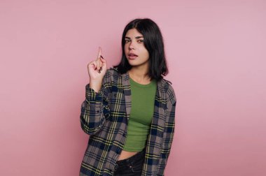 Genç bir kadın yeşil bluz ve ekose gömlek giymiş ve parmağını kaldırmış bir şekilde güvenle ayakta duruyor. Siyah saçları ve parlak pembe bir arkaplanı olan rahat bir yüzü var..