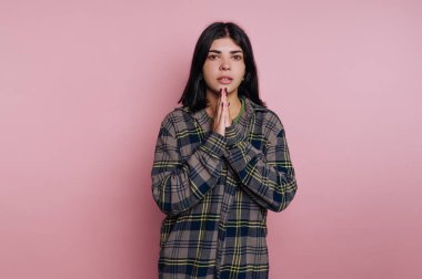 Genç bir kadın, elleri önünde sıkıca bağlanmış, ekose gömlek giymiş şekilde poz veriyor. Basit pembe arka plan onun ifadesini ve gelişigüzel tarzını vurgular..