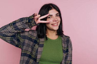 Genç bir kadın ekose gömlekle poz verirken özgüvenle gülümsüyor. Parmaklarıyla pembe bir arka plana yaslanmış bir barış işareti gösteriyor. Eğlenceli ve canlı bir atmosfer yaratıyor..