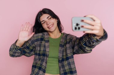 Genç bir kadın canlı pembe bir duvarın önünde duruyor, gülümsüyor ve selfie çekerken el sallıyor. Yeşil bir bluz ve kareli bir gömlek giyiyor, neşe saçıyor..