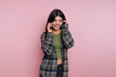 Genç bir kadın güvenli bir şekilde gülümsüyor ve telefonuyla konuşuyor, modaya uygun bir ekoseli gömlek ve yeşil bir bluz giyiyor, yumuşak pembe bir arka planda..