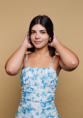 Çiçek desenli bir elbise giymiş genç bir kadın, elleri başının üstünde güvenle ayakta duruyor. Basit bej arkaplan onun tarzını ve duruşunu vurguluyor, çekici bir kompozisyon oluşturuyor.