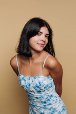 Genç bir kadın sırtı hafifçe dönük bir şekilde özgüvenle ayakta duruyor. Mavi çiçekli bir elbise giyiyor ve omuz hizasında koyu renk saçları var. Arkaplan sıcak bir bej rengi..